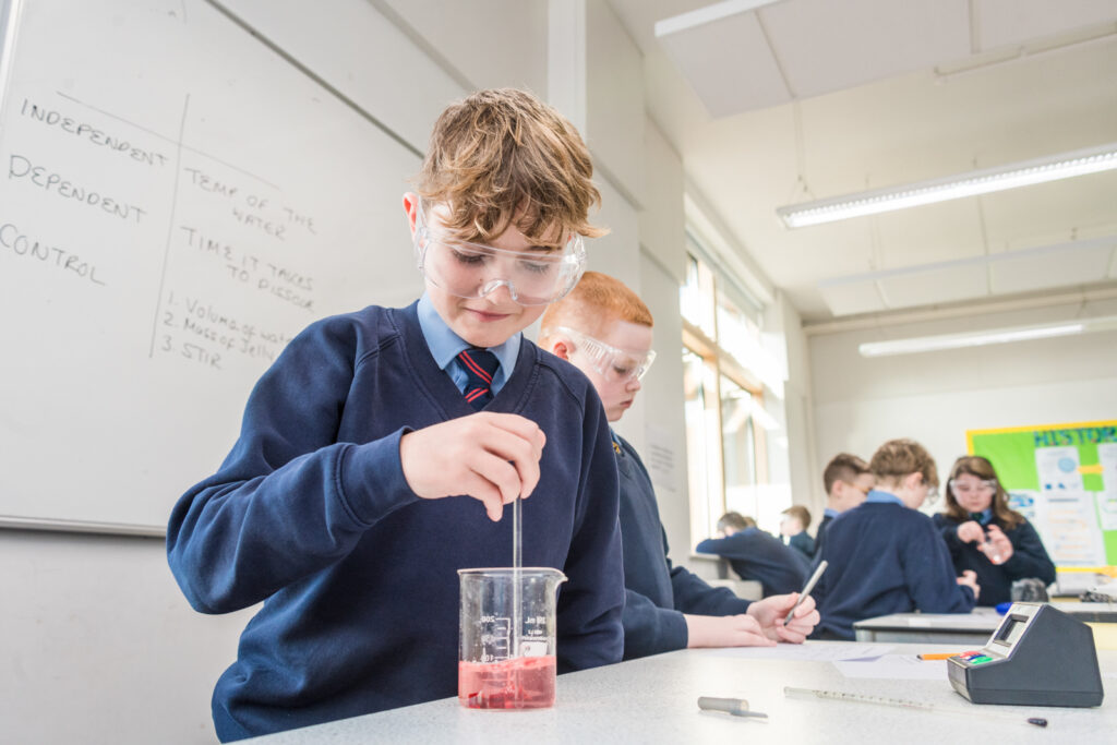 A male student wearing protective glasses and stirring a coloured liquid in a beaker, in a Science room